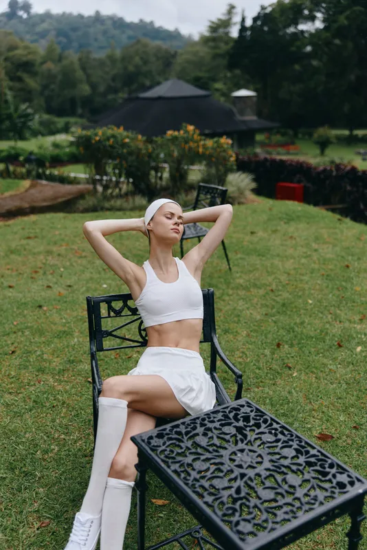 Stylish young model in a white crop top and skirt relaxing outdoors, showcasing a fit lifestyle amidst vibrant greenery, perfect for wellness and fitness themes