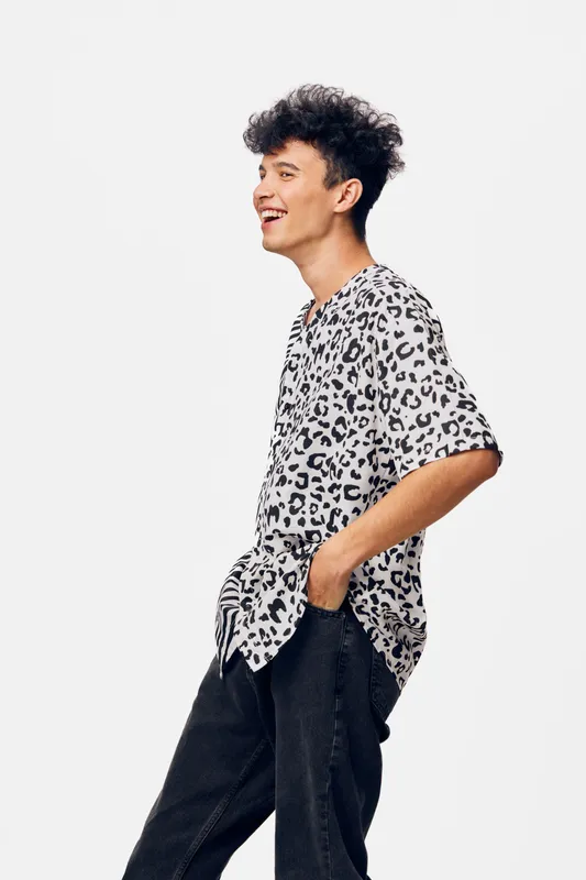 Young man wearing a casual leopard print shirt and black pants, smiling joyfully with hands in pockets against a plain white background.