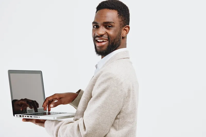 Smiling young man with dark skin and beard holding a laptop in hand, wearing a beige sweater, looking to camera, cheerful expression, casual clothing, isolated on white background, studio shot. People