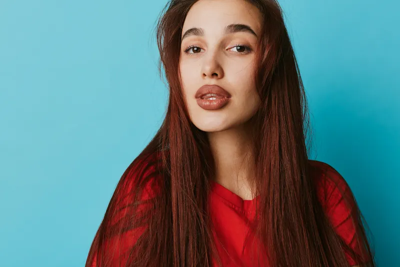 Young woman with long straight hair wearing red sweater posing on blue background. Portrait of female model with natural makeup and confident expression in studio lighting.