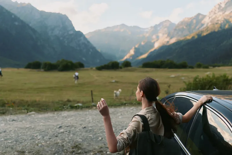 Woman traveler by a car in a mountain valley, gazing toward distant peaks, backpack ready, sunlight and open air, capturing a peaceful outdoor lifestyle moment