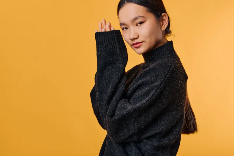 Young woman in dark sweater standing on orange background with hands near face looking confident and calm. Portrait of thoughtful female with long hair wearing casual warm clothes.