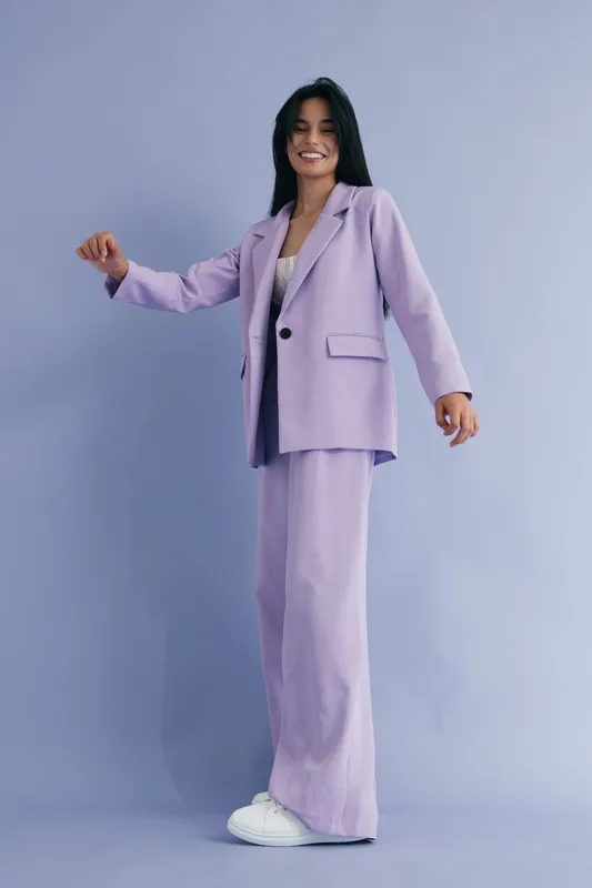 Young woman in lilac suit smiling confidently while standing against soft pastel blue background, wearing white sneakers and casual chic style.