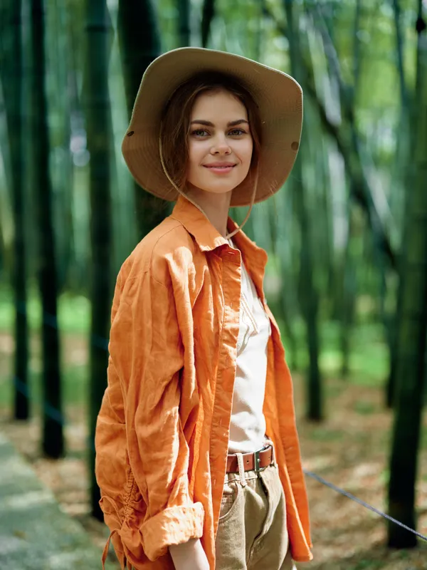 Woman in hat and orange jacket standing in bamboo forest, portrait with gentle smile outdoors. Casual travel style with natural sunlight, warm tones, relaxed mood and eco nature vibe.