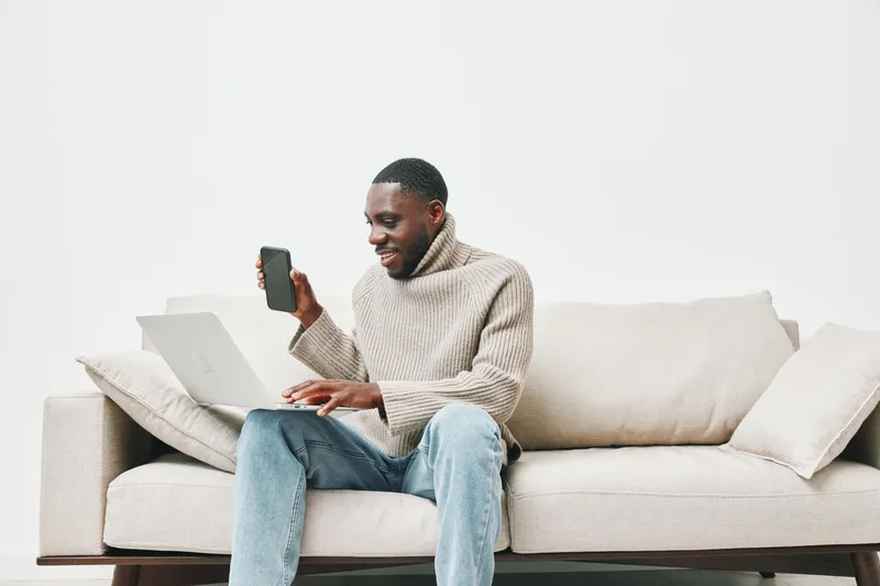 Smiling black man using laptop and smartphone on a cozy sofa, wearing a stylish sweater, showcasing a relaxed and modern lifestyle