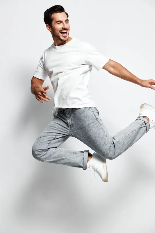 Happy cheerful young man jumping in the air wearing white t-shirt and patterned pants with joyful expression isolated on light gray background. Lifestyle concept.
