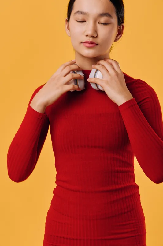 Young woman in red long sleeve dress holding white headphones close to neck posing with closed eyes. Studio portrait on orange background with calm expression and straight hairstyle.