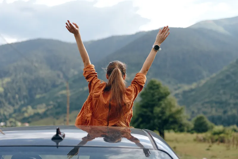 Woman standing on a car roof with arms raised in a scenic outdoor setting, mountains in the background, conveying freedom, celebration and a vibrant nature atmosphere.