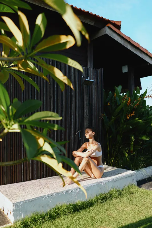 Relaxed woman in bikini sits on a concrete ledge beside a dark wooden wall, surrounded by tropical plants, enjoying sunny outdoor leisure and a casual lifestyle by a resort style retreat
