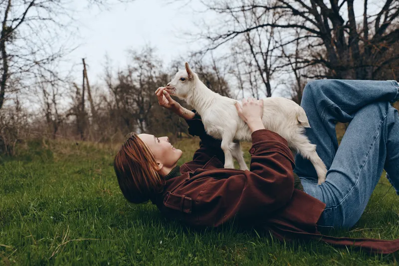 Relaxed woman lying on grass, playing with a goat kid, warm sunlight, gentle breeze, candid outdoor moment, countryside atmosphere, natural textures, soft colors.
