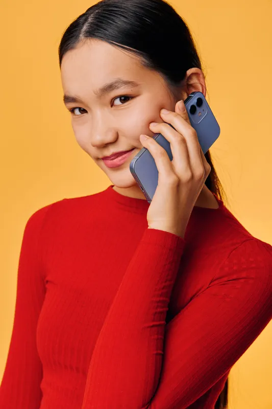 Young woman wearing red sweater holding smartphone near face smiling with confident expression on yellow background. Communication, technology, connection and modern lifestyle concept.
