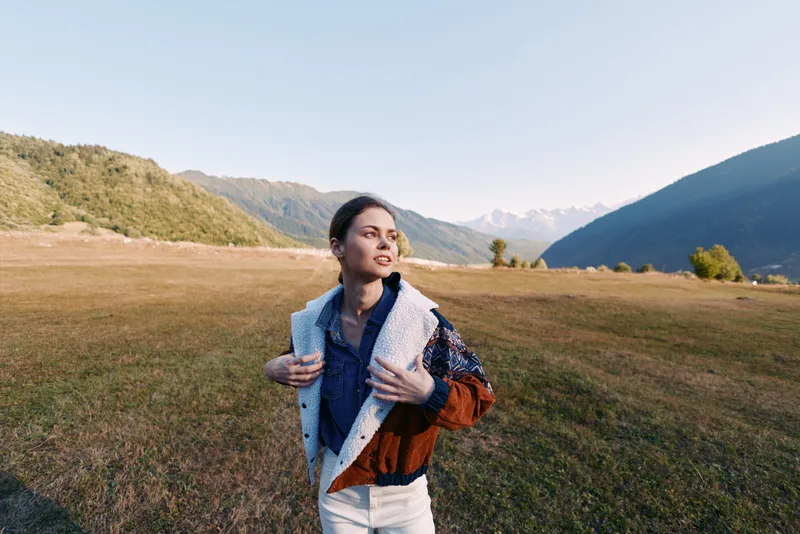 Woman in a shearling jacket stands in a wide field in a mountain valley, looking into the distance. Outdoor portrait with grass, hills and clear sky conveying travel, calm and adventure.