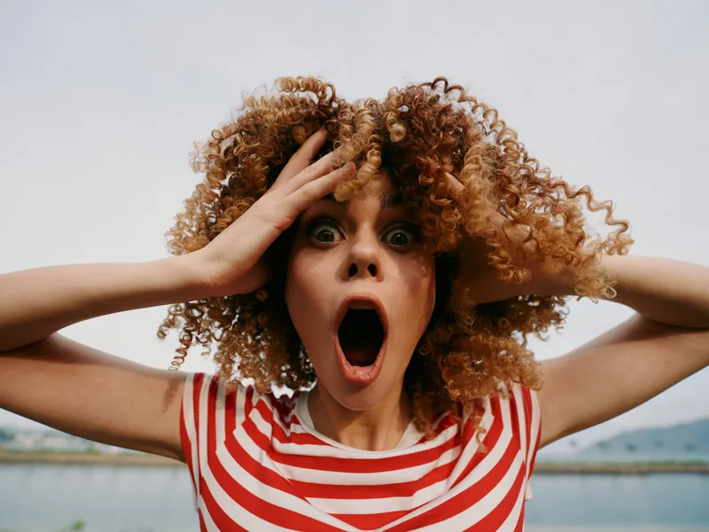 Close portrait of a surprised woman with vivid curly hair, hands on her head, mouth wide open, wearing a red and white striped shirt against a neutral backdrop.