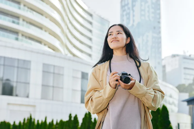Woman holding camera in outdoor urban setting, smiling amid modern buildings and glass facades, ready to capture candid street moments during daylight in a cityscape