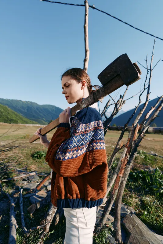 Woman axe wood countryside mountains sweater outdoor rural profile carrying large splitting axe over shoulder near wooden fence and stacked logs in mountain meadow landscape.