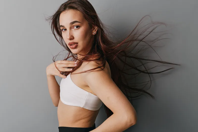 Young woman with long flowing hair and confident expression posing in white top against gray background. Female model with natural makeup and casual style looking at camera indoors.
