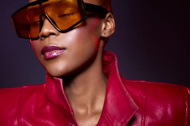 Close-up of confident black woman with dark skin wearing oversized orange sunglasses and red leather jacket. Studio portrait with purple background. Concept for fashion, style, and attitude.