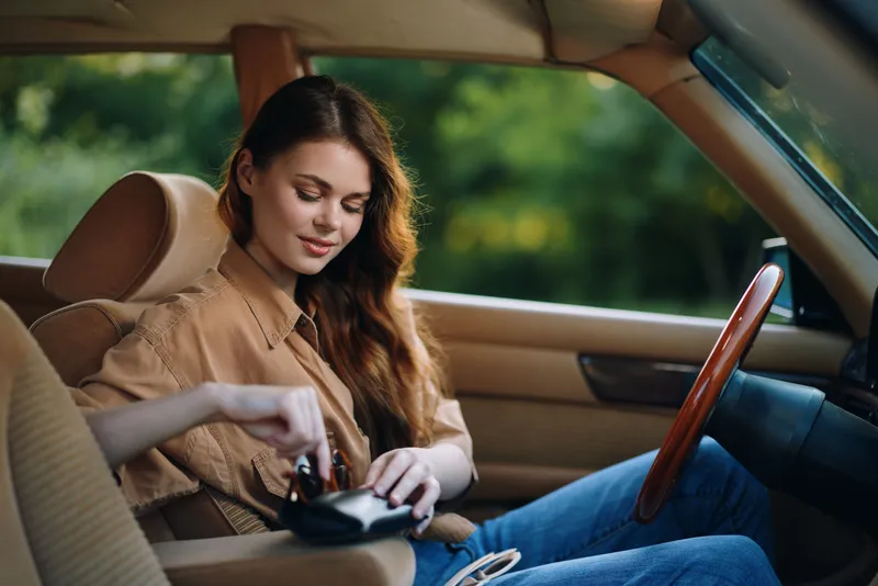Young woman in casual attire with long hair, sitting in a car, smiling while organizing her belongings, surrounded by green nature, showcasing a relaxed summer vibe.