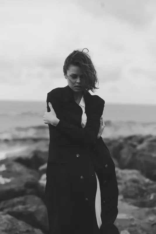 Woman wearing a long black coat stands on a rocky coast, wind sweeping her hair, moody monochrome scene, fashion and solitude by the sea, dramatic urban vibe meets rugged nature, cinematic
