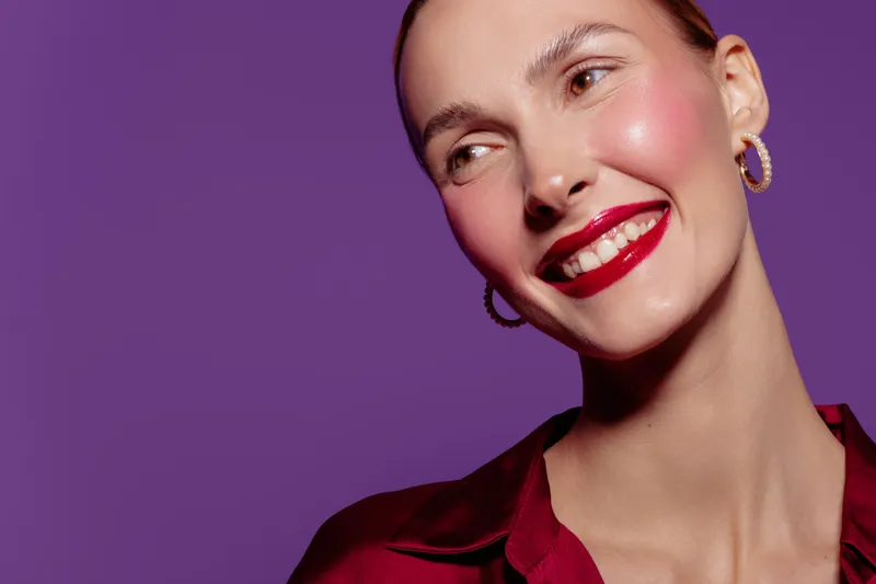 Smiling woman with red lipstick and hoop earrings looks away on purple background. Close-up portrait with shiny skin and red satin blouse. Concept of beauty, fashion, and confident mood.