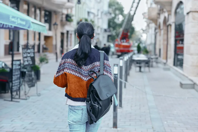 Backpack, street, walking, urban fashion, traveler, city scenery and casual outfit on an individual walking away along a shop lined avenue in morning light