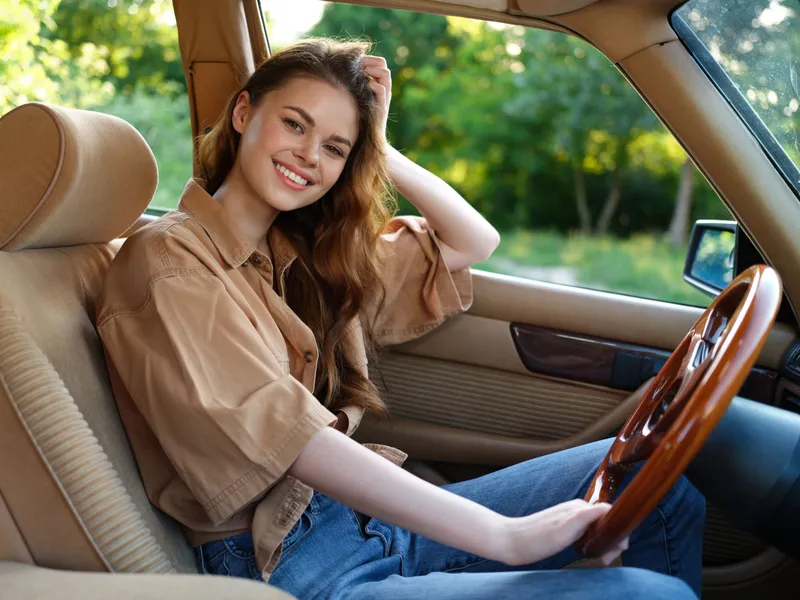 Young woman smiling in a vintage car, wearing a casual brown outfit, with sunlight filtering through green trees, evoking a sense of freedom and joy.