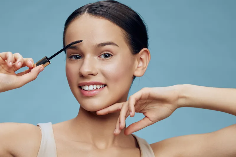 Young woman applying mascara with a smile against a soft blue background, showcasing a natural beauty look ideal for makeup promotions and beauty campaigns