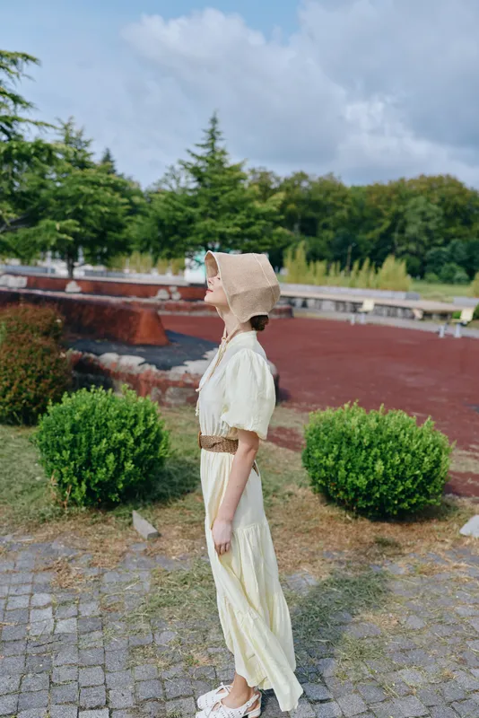 Elegant woman in a cream vintage dress and wide hat strolls along a sunny park path, creating a nostalgic fashion moment in a timeless outdoor setting.