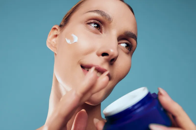 Happy woman applies moisturizer from a jar, smiling as cream sits on her cheek, isolated on a blue background, showcasing a clear skincare routine and healthy glowing skin. Daily.