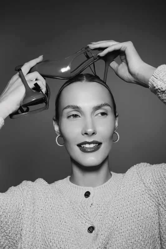 Young white woman with fair skin holds high heel shoe above head, smiling softly. Concept of fashion, beauty, and elegance in studio portrait.