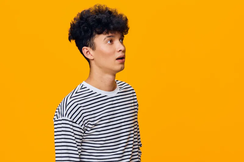 surprised man with curly hair wearing striped shirt stands against bright orange background, expressing shock and curiosity, youthful and casual style portrait.