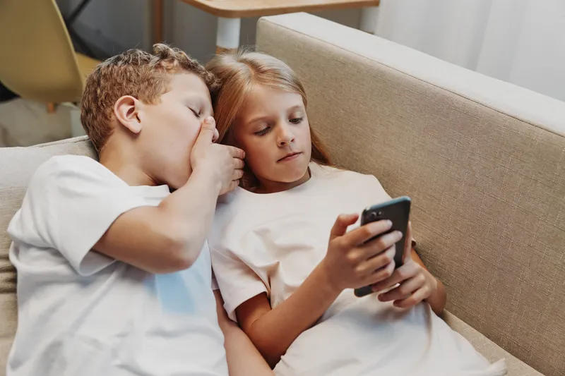 Two children sit on a beige sofa indoors, one boy whispering a secret to a girl holding a smartphone. Casual moment of friendship and communication in a cozy home setting.