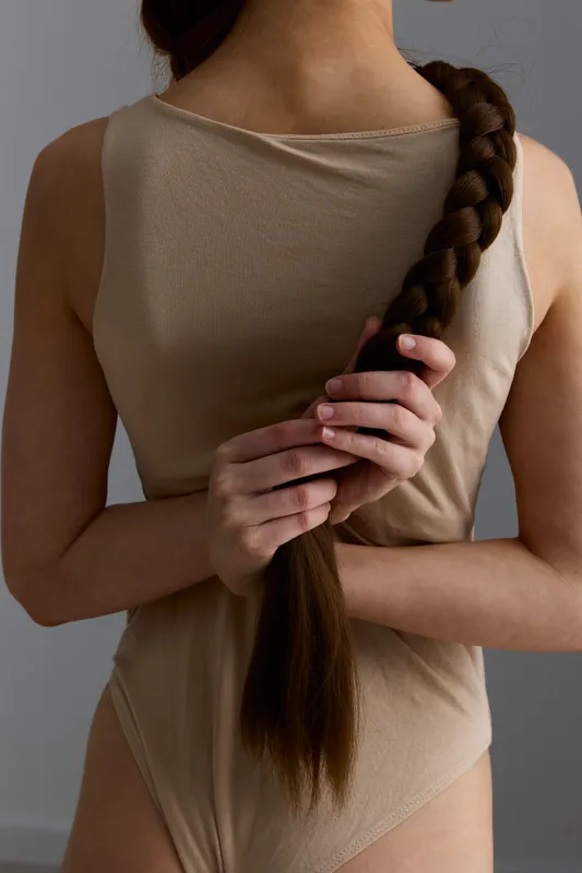 Woman with long braided hair holding her braid behind back, wearing a beige sleeveless top and bodysuit against neutral gray background in soft natural light.