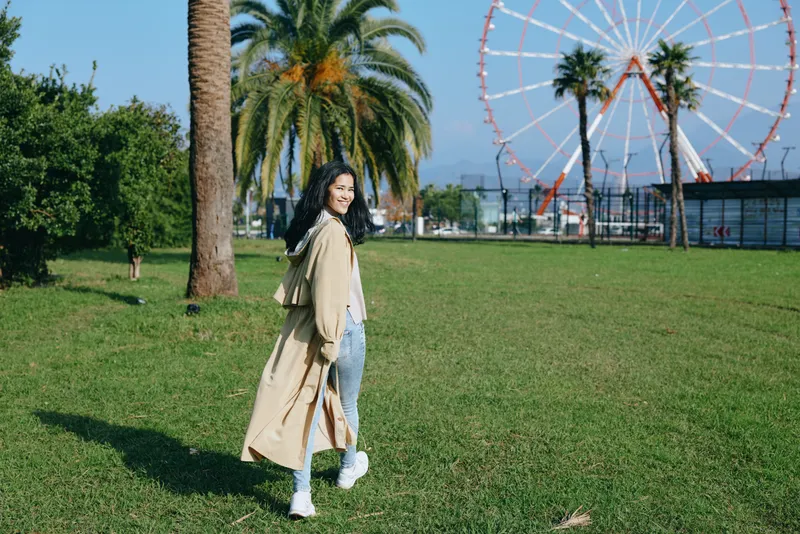 Woman in a beige coat strolling on a sunny park lawn with a ferris wheel in the background, casual outdoor leisure, travel vibe and fresh air.