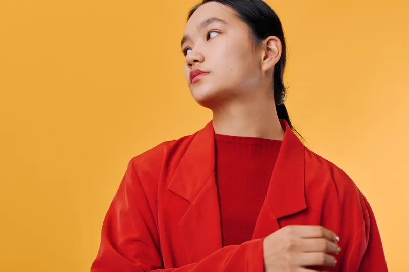 Young woman in red jacket looking to side on yellow background, portrait of thoughtful female with dark hair tied back wearing casual bright clothes in studio setting.
