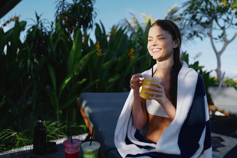 A young woman sits poolside with a towel over her shoulders, smiling as she drinks a bright yellow beverage, enjoying a sunny outdoor moment by lush green plants.