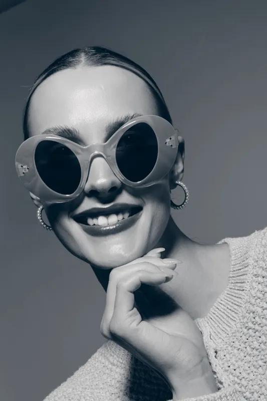 Young Caucasian woman with fair skin wearing large round sunglasses and hoop earrings smiles confidently. Fashion portrait with stylish accessories and casual sweater.