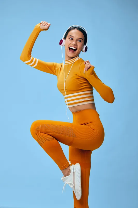 Fitness woman in bright orange athletic wear dances with headphones, showcasing energy, movement and vibrant sport vibe against a solid blue backdrop, capturing joy and active lifestyle.