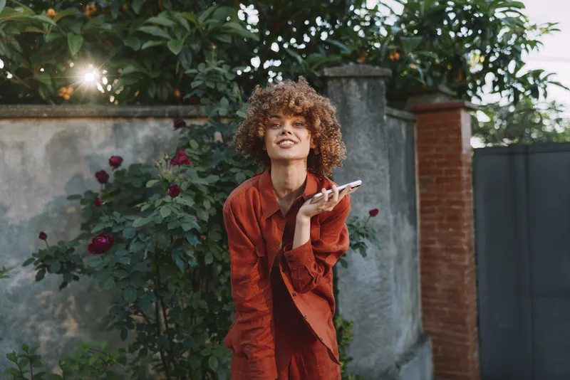 A smiling woman in a red coat poses outdoors in a garden, holding a phone. A candid lifestyle portrait with sunshine, urban vibe, and a cheerful mood, showcasing confidence and style