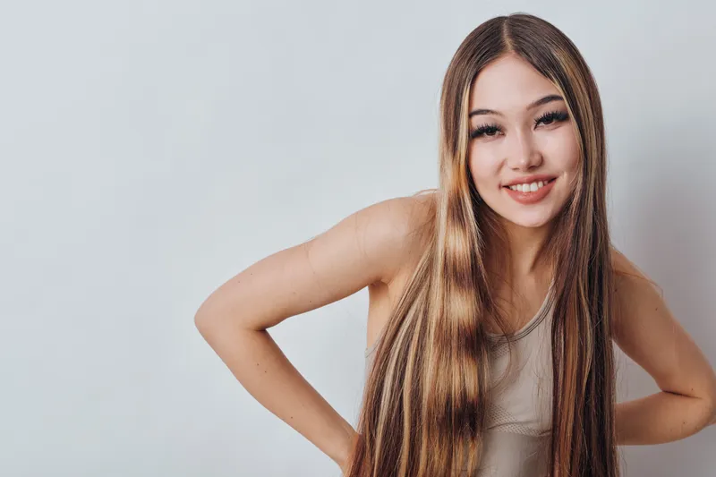 Young woman with long straight hair smiling with hands on hips, wearing casual sleeveless top against gray background in studio portrait. Positive expression and confident pose.