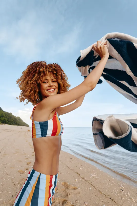 Happy young woman with curly hair wearing colorful striped swimsuit holds towel on sandy beach near calm sea under blue sky with clouds, enjoying summer day outdoors.