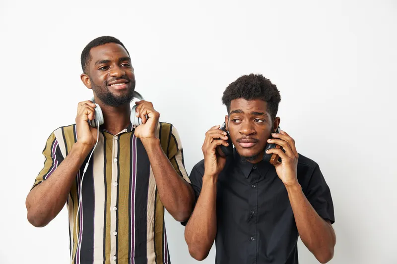 Two black men enjoying music together, one smiling with headphones while the other listens intently. Perfect for showcasing friendship and shared experiences.