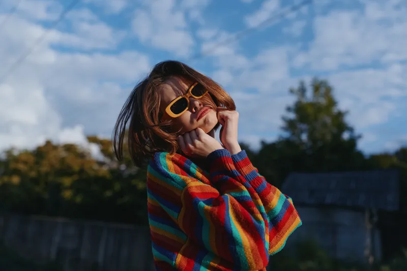 Young woman in her twenties wearing a colorful striped sweater and yellow sunglasses outdoors, smiling and posing with chin on hands, casual outfit, fashion lifestyle portrait.