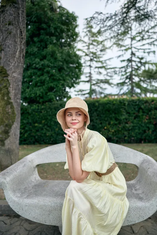 woman outdoor bench dress hat sitting on a curved concrete bench in a garden, smiling softly as sunlight lights her relaxed pose