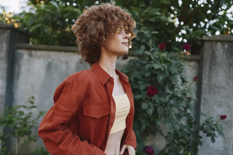 A confident woman with curly hair stands outdoors in a garden, wearing a rust shirt over a beige top. Bright natural light highlights her relaxed pose, capturing a lifestyle moment of self expression