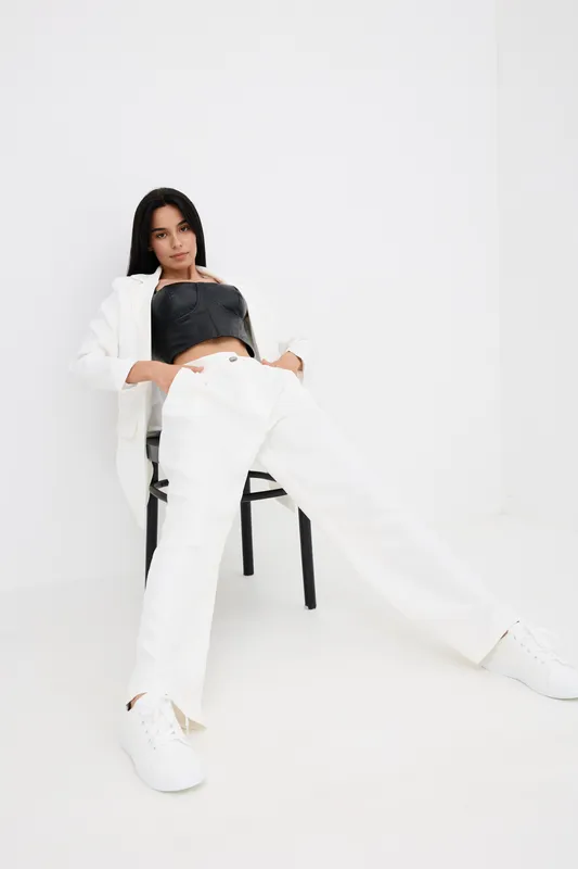 Young woman in white pants and jacket wearing black top, posing casually sitting on chair in minimalistic white room, modern fashion style, relaxed mood.