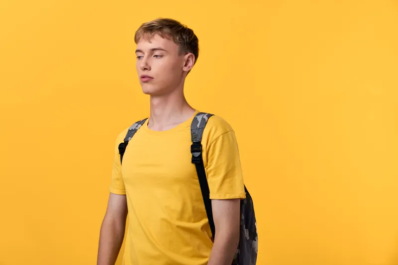 Youthful man in a yellow t shirt with a backpack stands before a bright background, exuding casual style, urban lifestyle, and readiness for travel or daily exploration