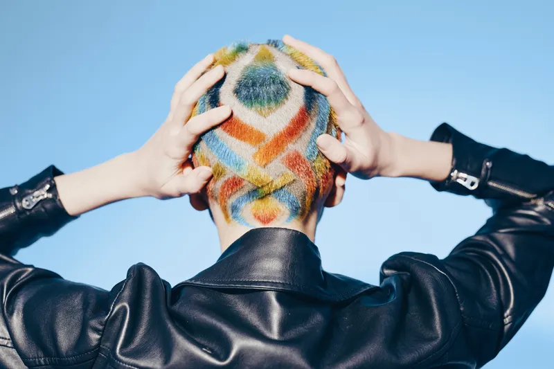 Woman with short hair dyed in colorful geometric patterns wearing a black leather jacket against a clear blue sky, showcasing edgy fashion and creative hairstyle.