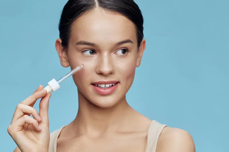 Young girl with light skin holding a skincare serum, smiling against a blue background, embodying a fresh and vibrant skincare routine