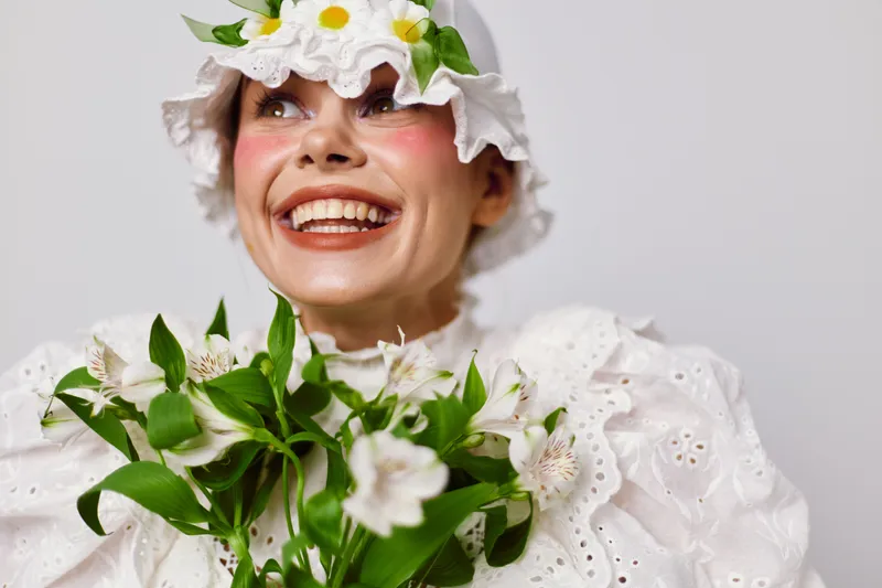 Cheerful model in a white floral dress holding flowers, embodying joy and spring freshness in a bright, minimalist setting